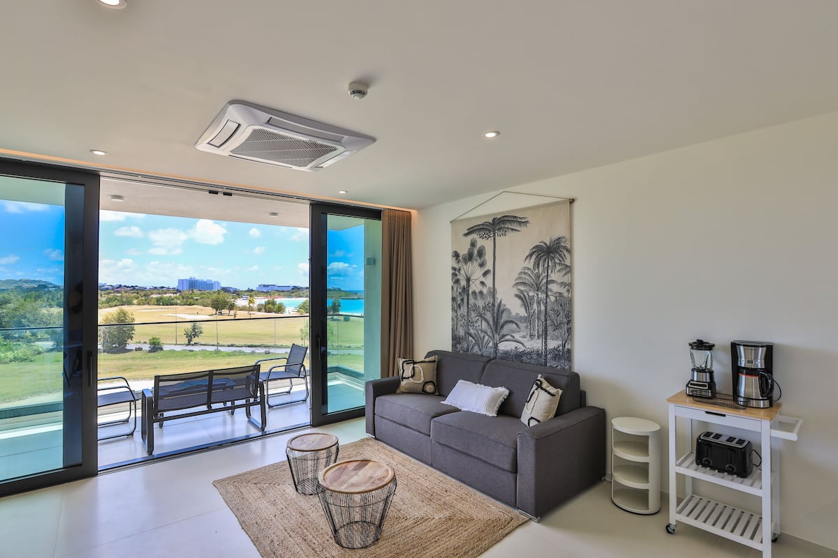Un salon moderne avec un canapé gris, un coin café et une vue sur un terrain de golf, visible à travers de grandes portes vitrées.