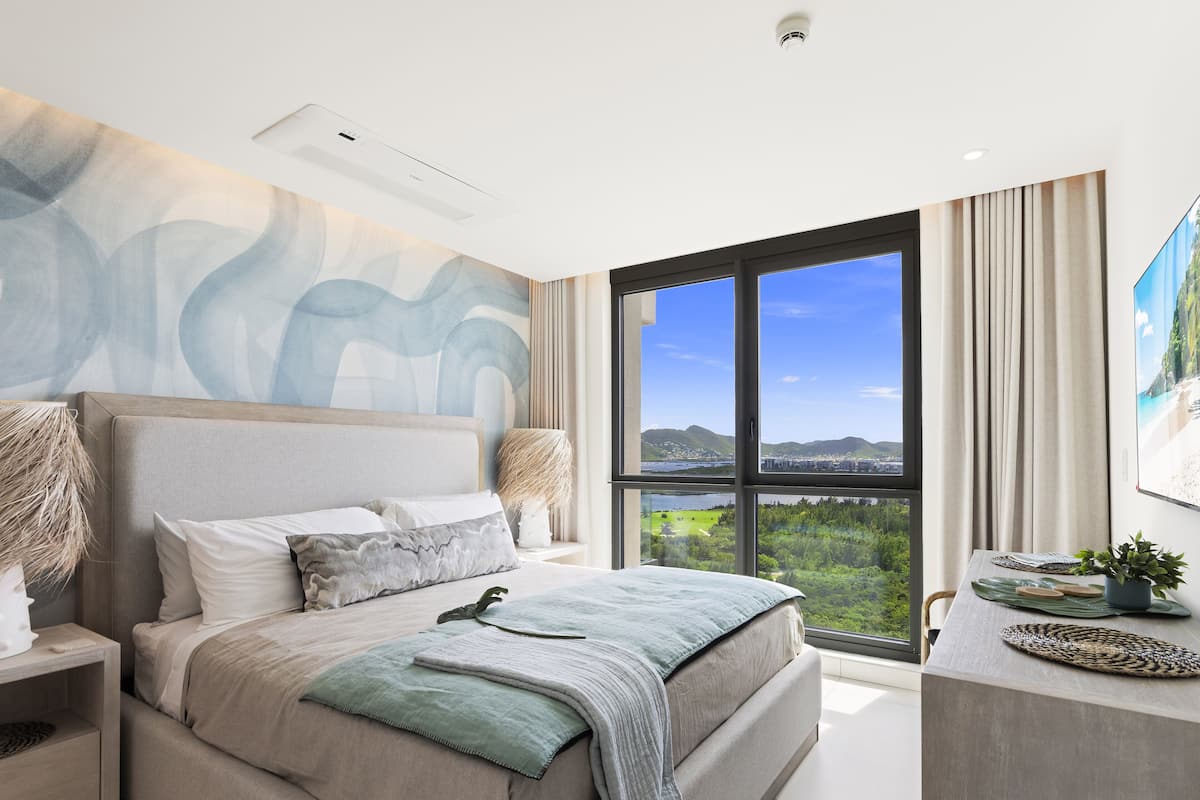 Une chambre moderne avec un grand lit, une table de chevet, un bureau et une vue sur un paysage côtier grâce à de grandes fenêtres.