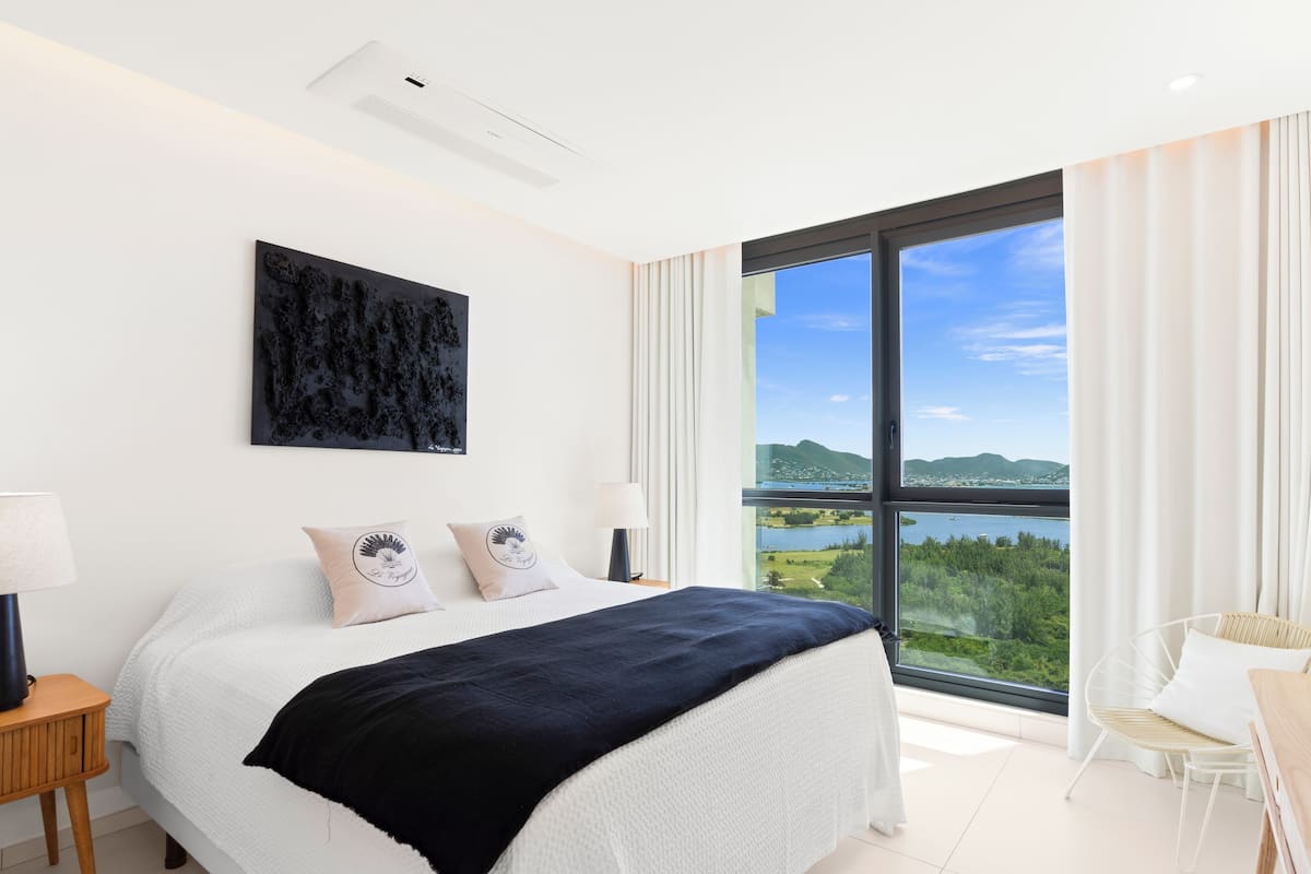 Une chambre moderne avec un grand lit, une vue sur un lac et des montagnes, et un paysage urbain visible au loin.