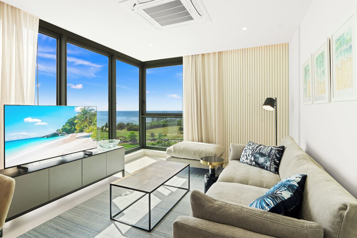 Un salon moderne doté d’un grand téléviseur, de sièges confortables, d’une table basse et offrant une vue sur une plage et une végétation luxuriante.