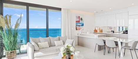 A modern living room with a large window overlooking the ocean, a dining area, and a kitchen with white cabinetry.