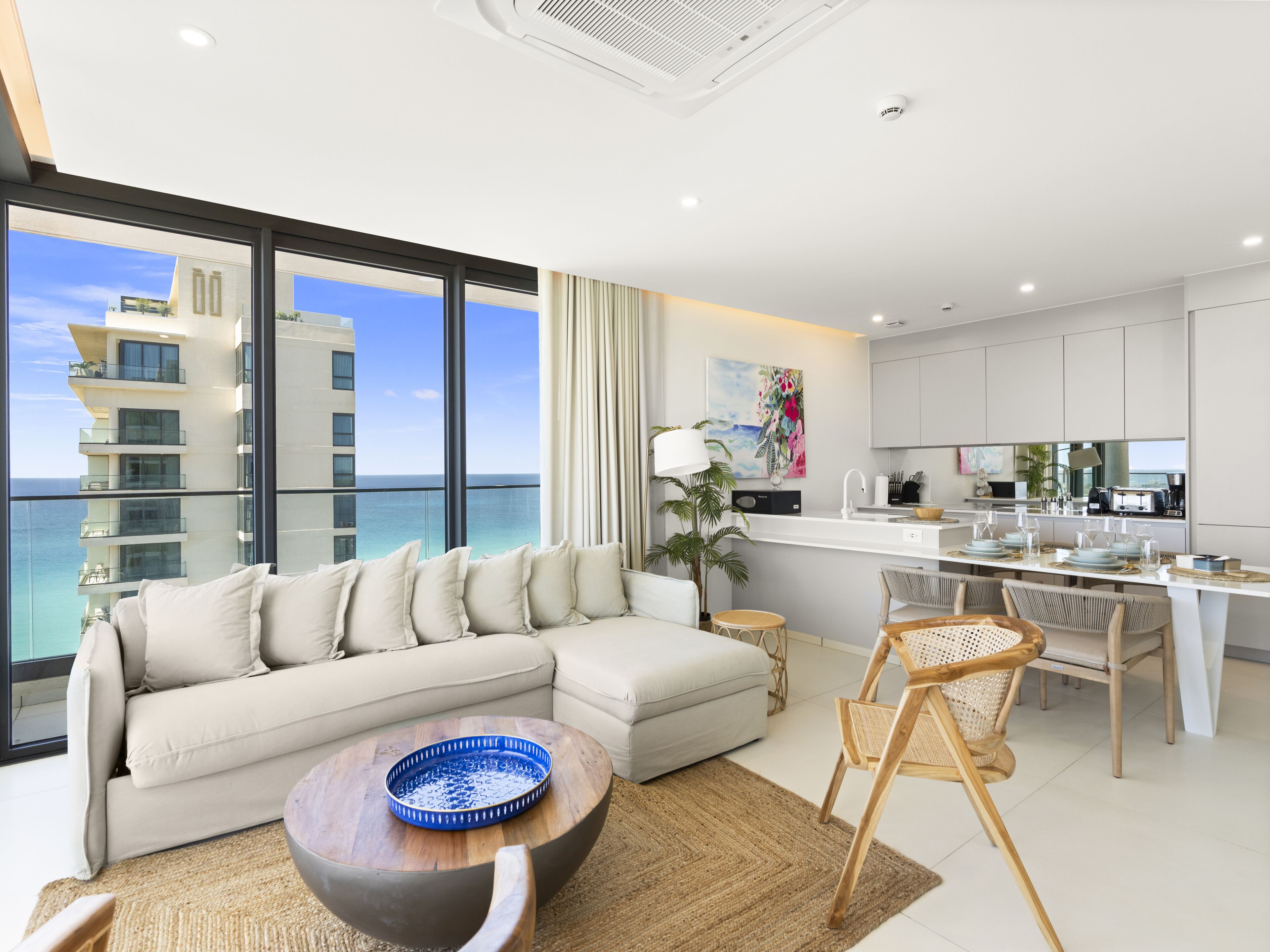A modern living room with a sofa, coffee table, and dining area, overlooking a balcony with a view of the ocean and buildings.