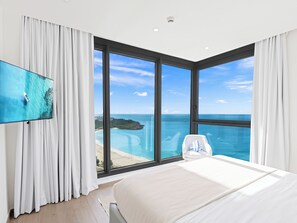 Une chambre d’hôtel moderne dotée d’une grande fenêtre donnant sur la mer, d’un téléviseur à écran plat fixé au mur et d’un lit blanc.