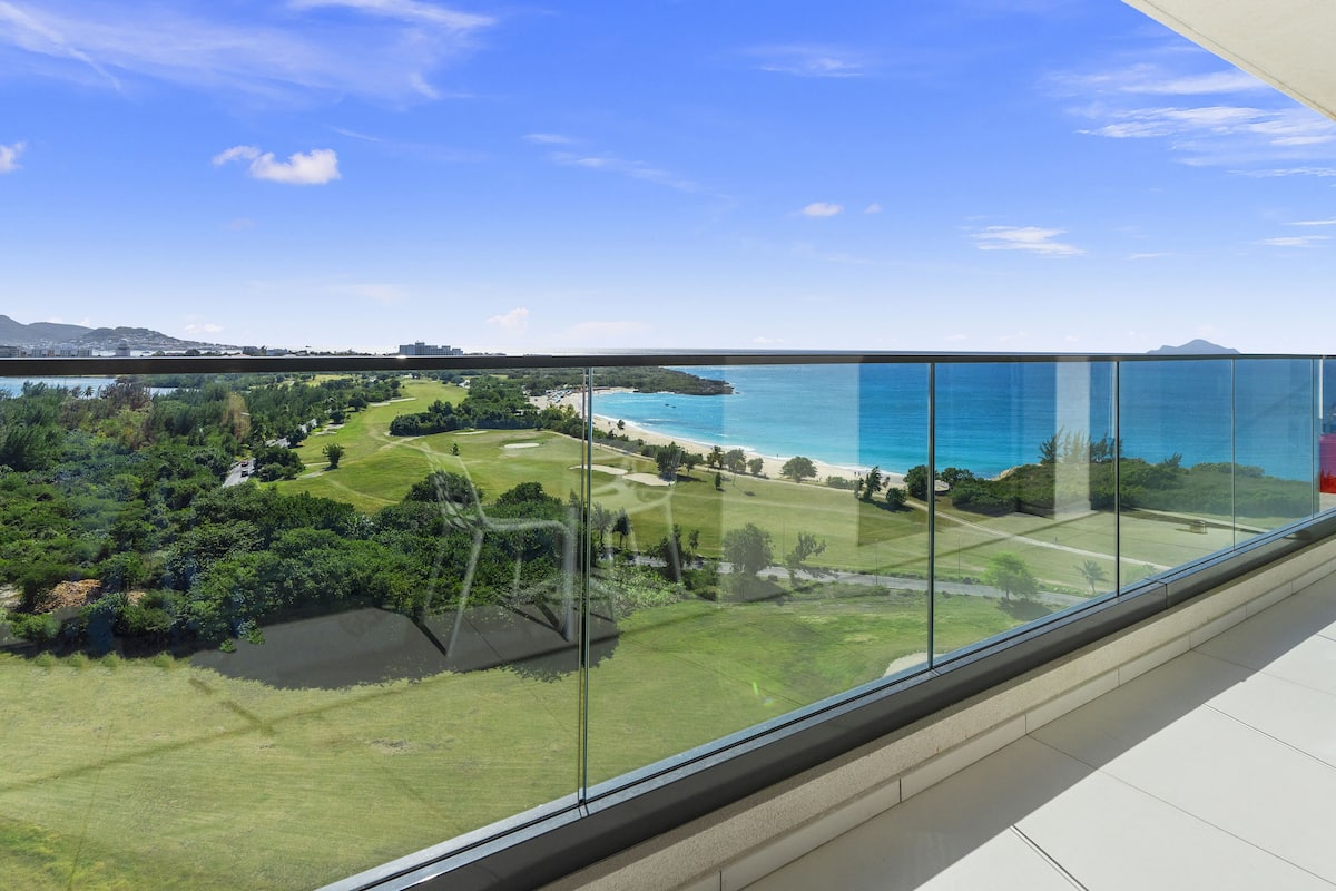 Un immeuble de grande hauteur doté d’un balcon en verre offrant une vue sur un paysage côtier, comprenant un terrain de golf et une plage.