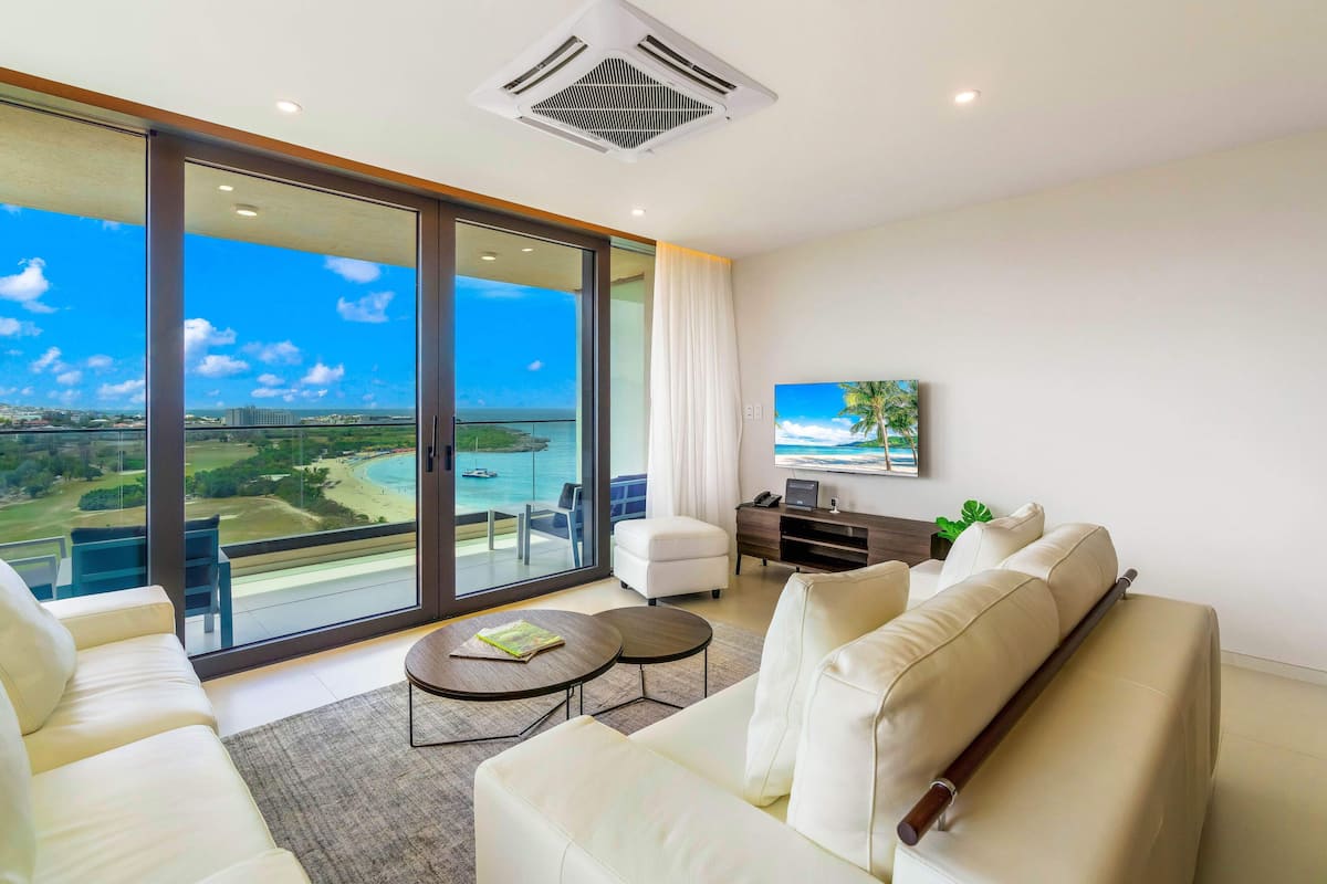 Un salon moderne doté d’une grande porte-fenêtre coulissante donnant sur un balcon avec vue sur la mer et un terrain de golf. On y trouve un canapé blanc, une petite table ronde et un téléviseur fixé au mur.