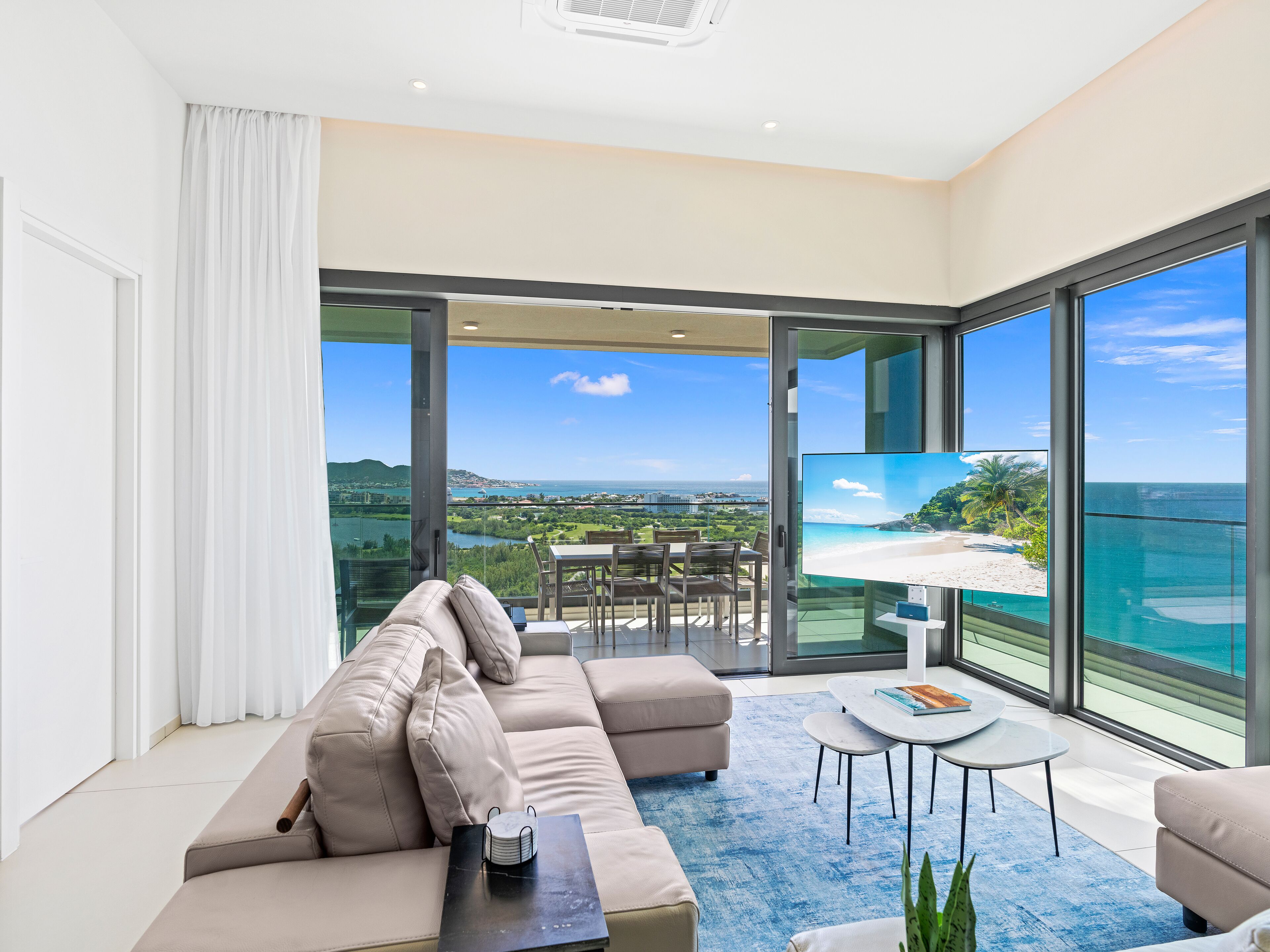 A modern living room with a large TV, comfortable seating, and a balcony with a scenic ocean view.