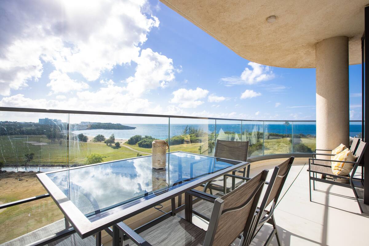 Un balcon doté d’une table et de chaises en verre, offrant une vue panoramique sur un paysage pittoresque comprenant une étendue d’eau et de la verdure.