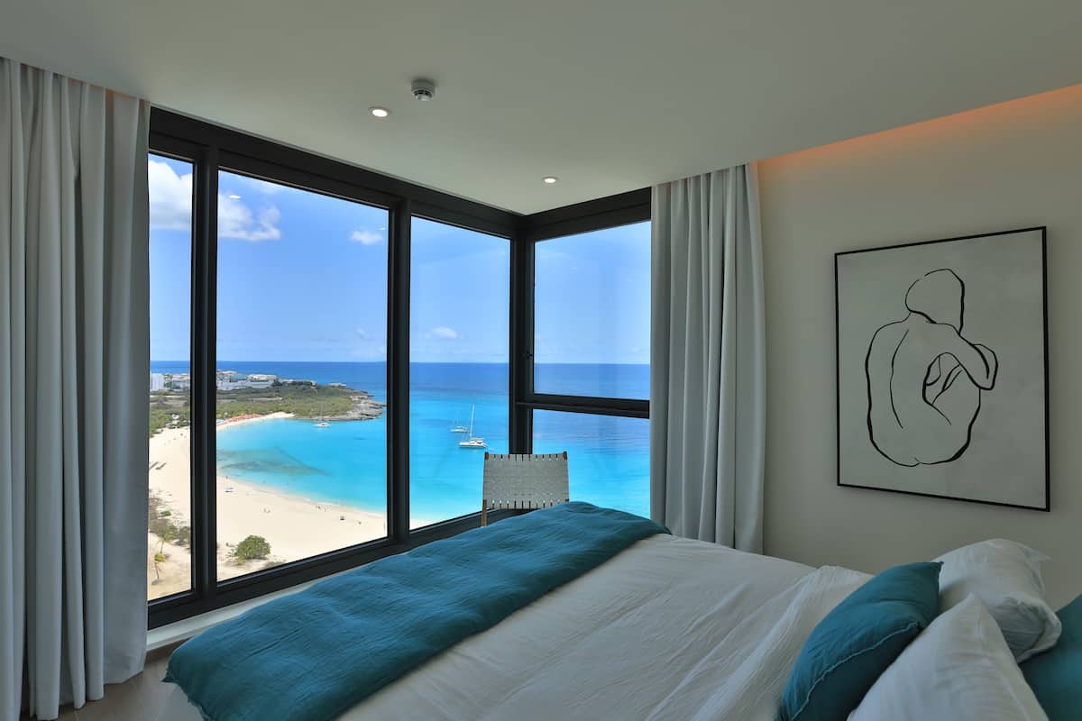 Une chambre avec une grande fenêtre donnant sur une plage et l’océan, un lit avec une literie blanche et turquoise, et une œuvre d’art encadrée accrochée au mur.