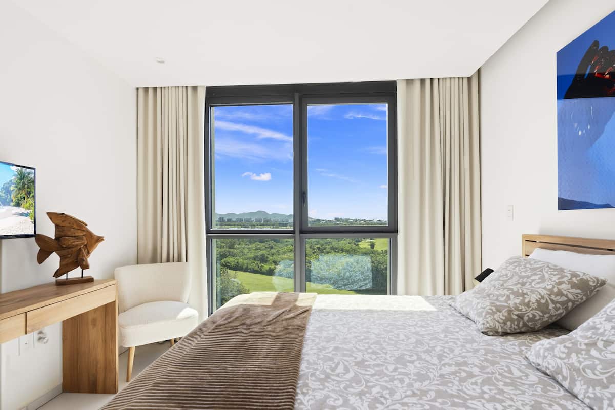 Une chambre d’hôtel moderne dotée d’un grand lit, d’un bureau avec un ordinateur et offrant une vue sur la plage et les montagnes par la fenêtre.