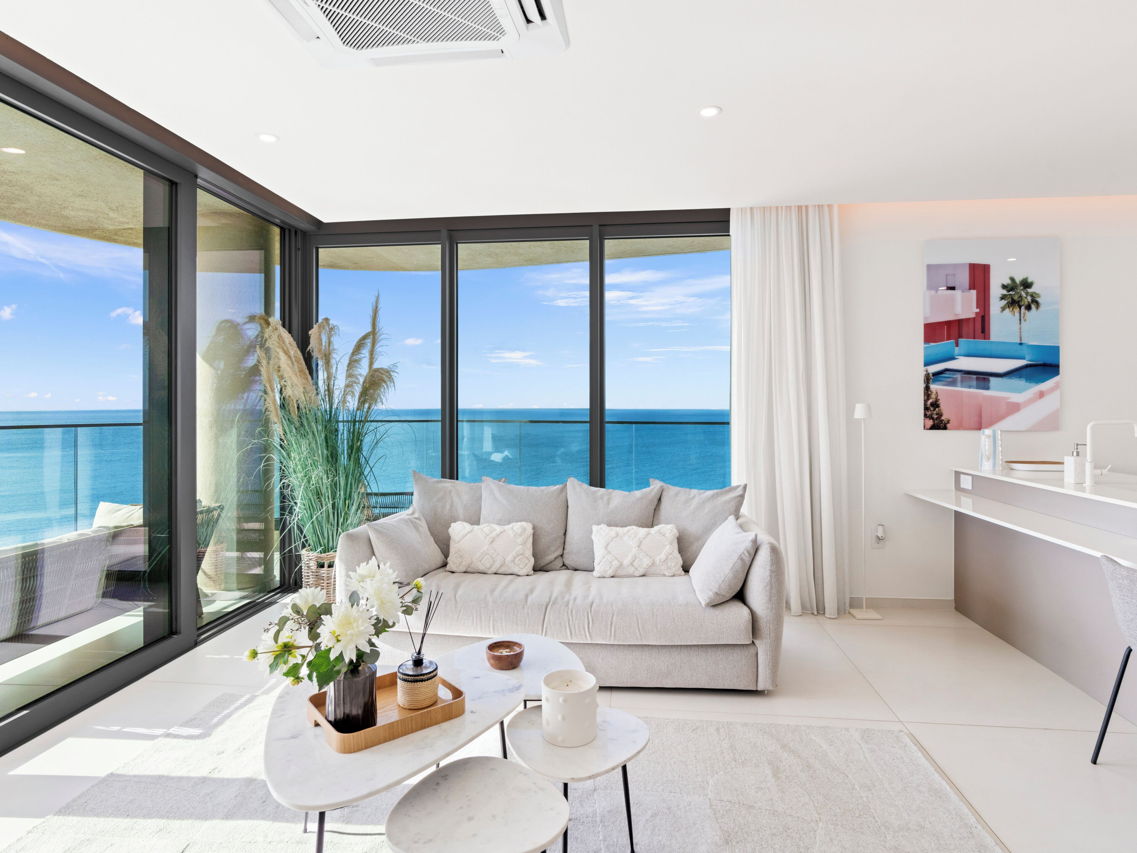 Un salon moderne doté d’une grande fenêtre donnant sur la mer, d’un canapé avec des coussins, d’une petite table sur laquelle est posé un vase de fleurs, et d’une salle de bain minimaliste avec un lavabo et un plan de toilette blancs.