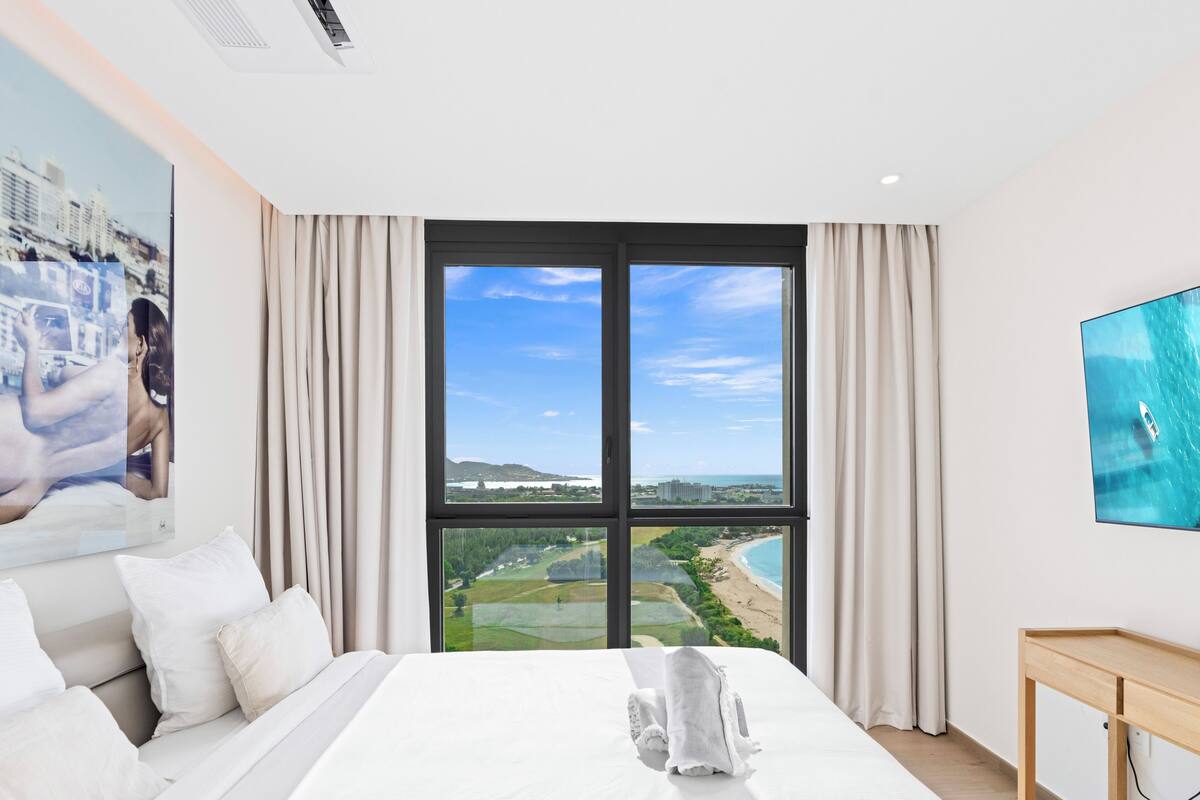 Une chambre d’hôtel avec un grand lit, un téléviseur fixé au mur et une vue sur la plage et le paysage urbain, visible par la fenêtre.