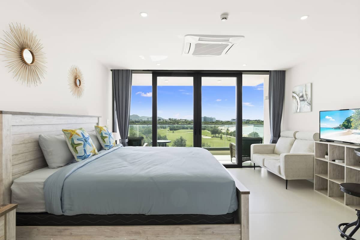 Une chambre d’hôtel moderne dotée d’un grand lit, d’un bureau avec un téléviseur à écran plat et d’un balcon offrant une vue sur un terrain de golf et une plage.