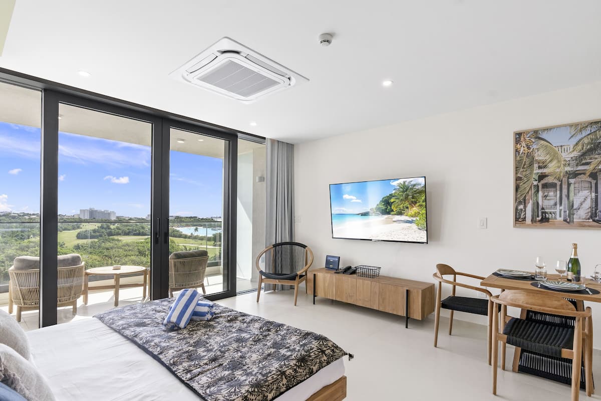 Une chambre d’hôtel moderne dotée d’un grand lit, d’un bureau avec un ordinateur, d’un coin repas et d’un téléviseur diffusant des images d’une plage.
