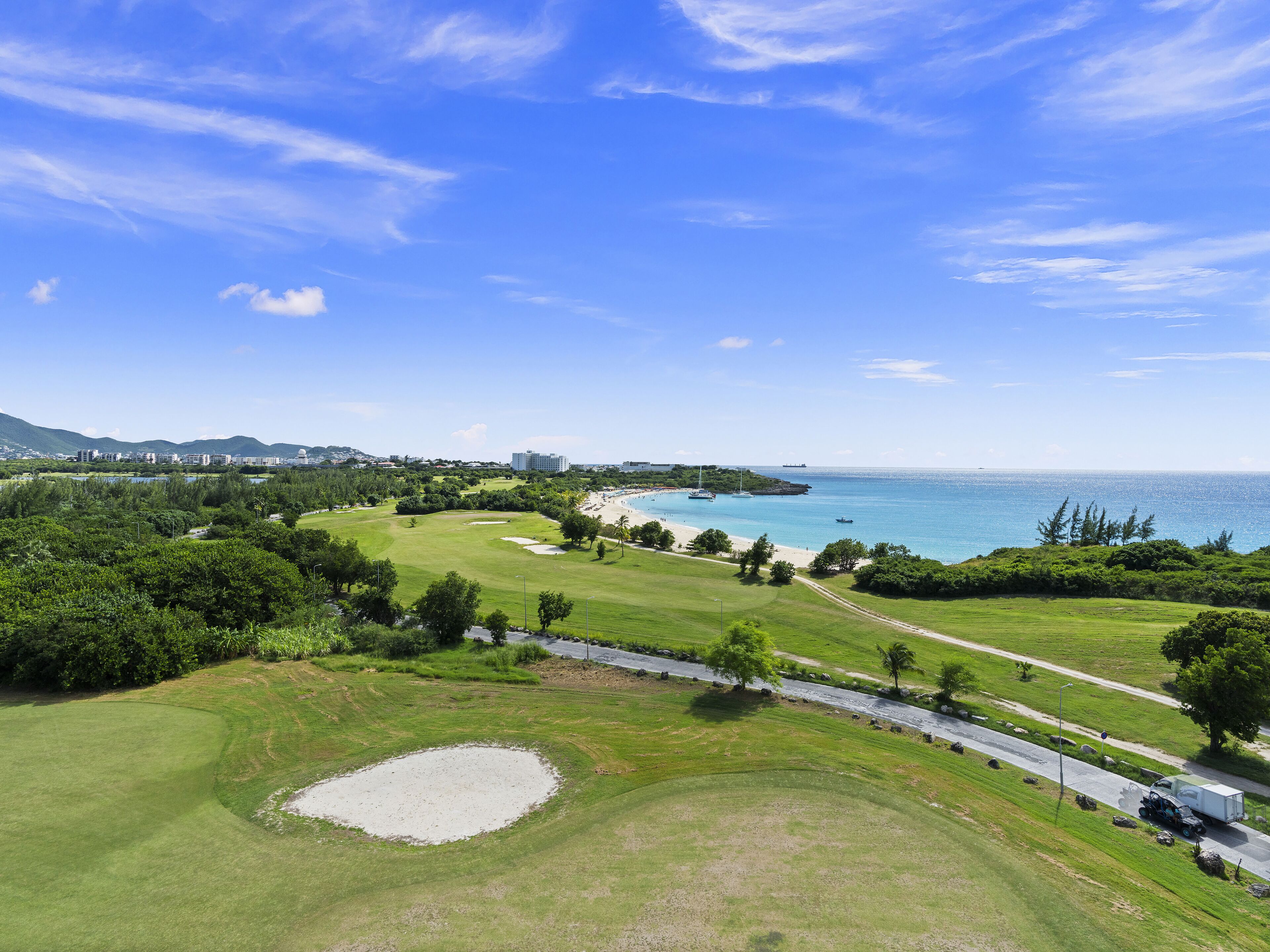 Um campo de golfe com uma grande área de areia, uma estrada com um caminhão e vista para o oceano e edifícios distantes.