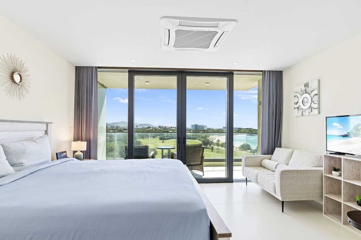 Une chambre d’hôtel moderne dotée d’un grand lit, d’un canapé, d’une télévision et d’un balcon offrant une vue sur la plage et les montagnes.