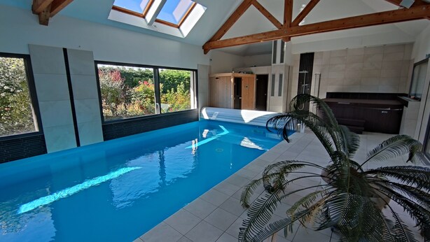 An indoor swimming pool with a view of the garden, wooden beams, and a sauna area.