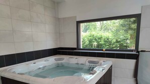 A modern bathroom with a large bathtub, a window with a view of greenery, and a wooden bench.