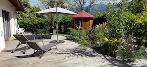 Een terras buiten met een tafel en stoelen, een houten prieel en een tuin met bloeiende planten.