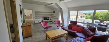 Un salón amplio con una mesa de centro de madera, un sofá de cuero rojo y dos sillones rojos. Un televisor está montado en la pared y se aprecia una vista al exterior a través de amplios ventanales.
