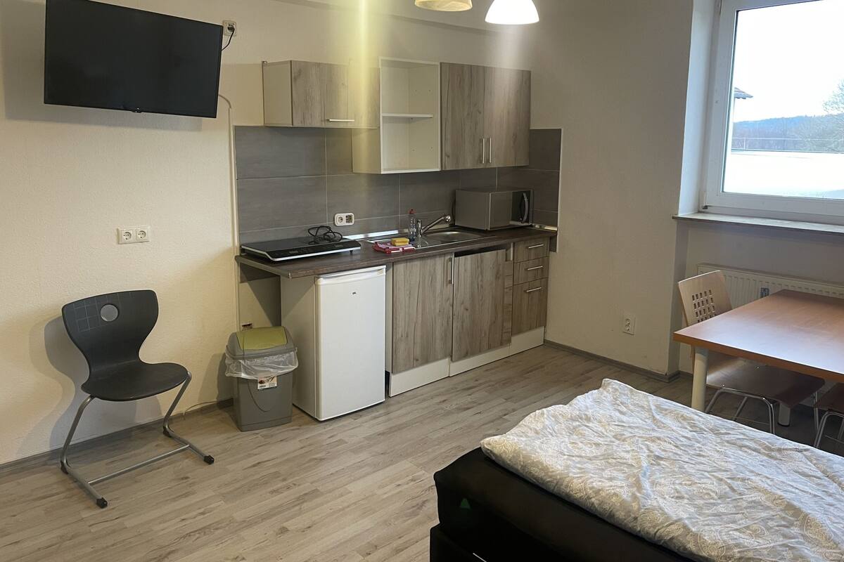 Eine kompakte Küchenzeile mit Mikrowelle, Spüle und Schränken, ein kleiner Esstisch mit Stühlen, ein Bett mit gemustertem Duvet und ein an der Wand befestigter Fernseher.