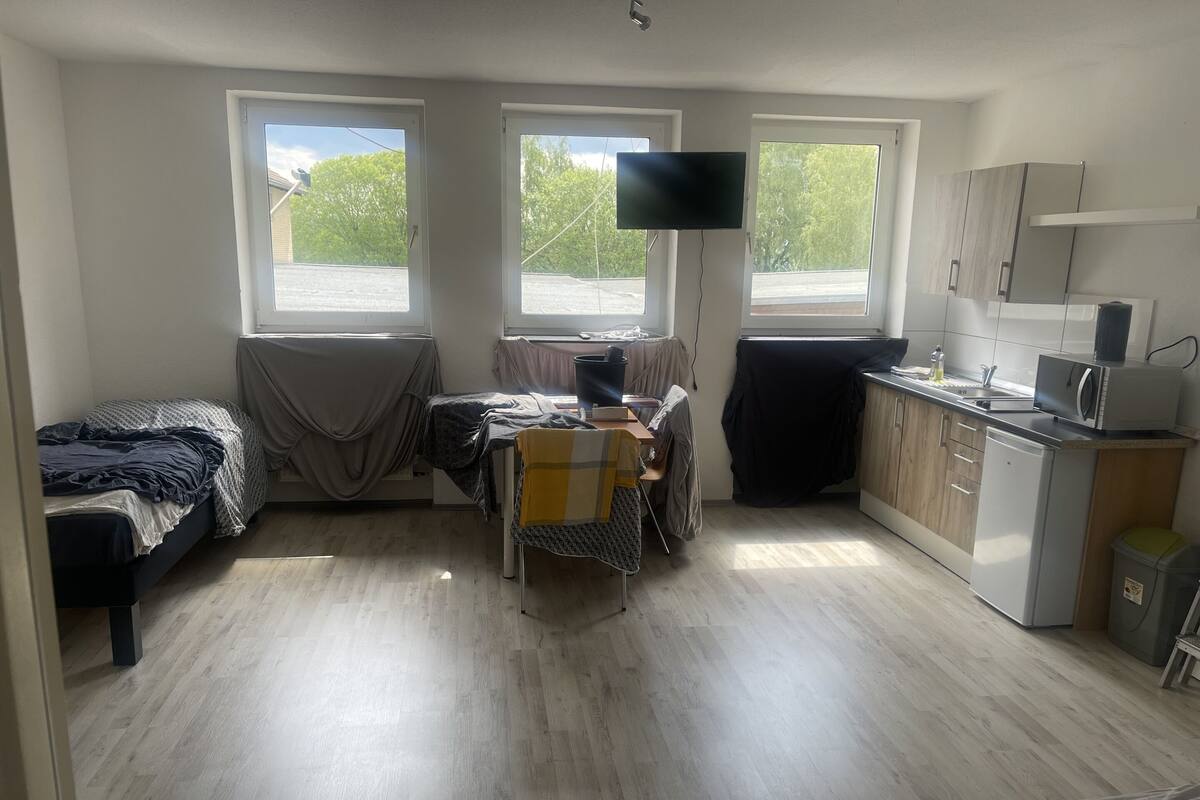Ein kleines Zimmer mit Bett, einem Tisch mit Stuhl, einem an der Wand befestigten Fernseher und einer Küchenzeile.