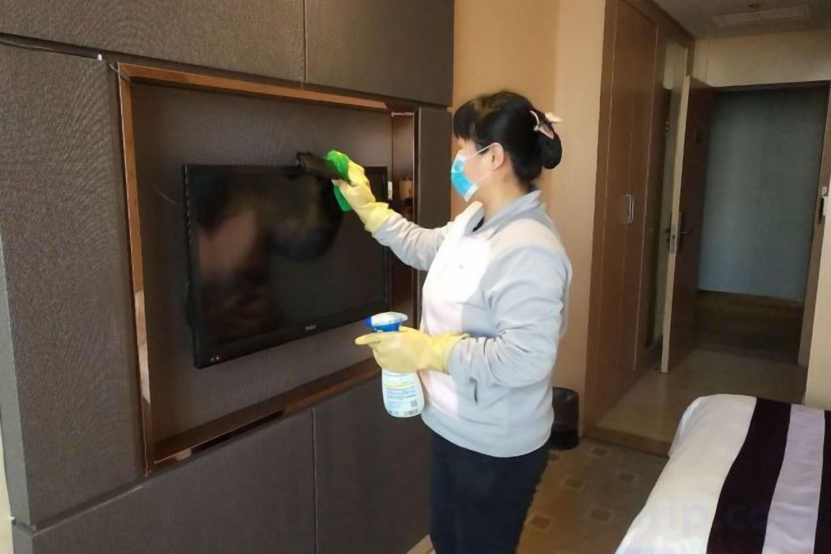 Uma pessoa limpando uma TV de tela plana fixada na parede de um quarto, com uma cama e uma porta ao fundo.