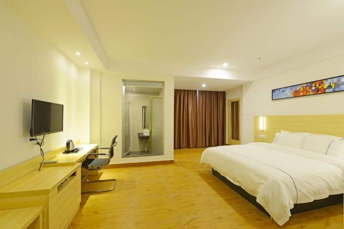 A hotel room with a bed, a desk with a chair, a television, a shower area, and a painting on the wall.