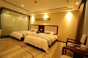 Habitación de hotel con dos camas, cabina de ducha de vidrio, mesita de noche con lámpara y una pequeña zona de estar con una silla y una mesa.