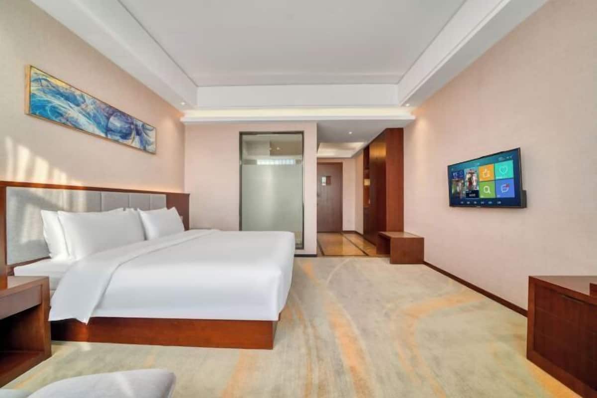 A hotel room with a bed, a TV, a painting, and a hallway leading to another room.