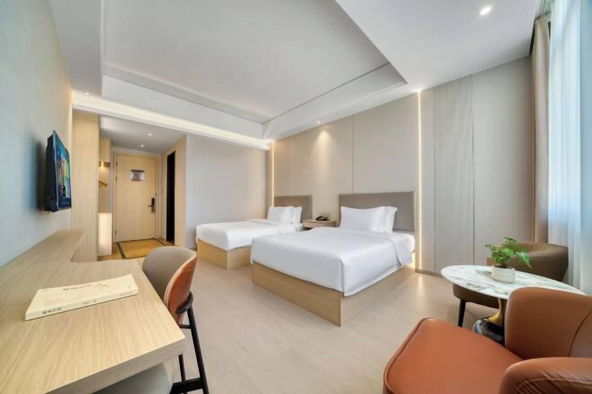 A hotel room with two beds, a desk, a chair, a small table with a plant, and a TV on the wall.