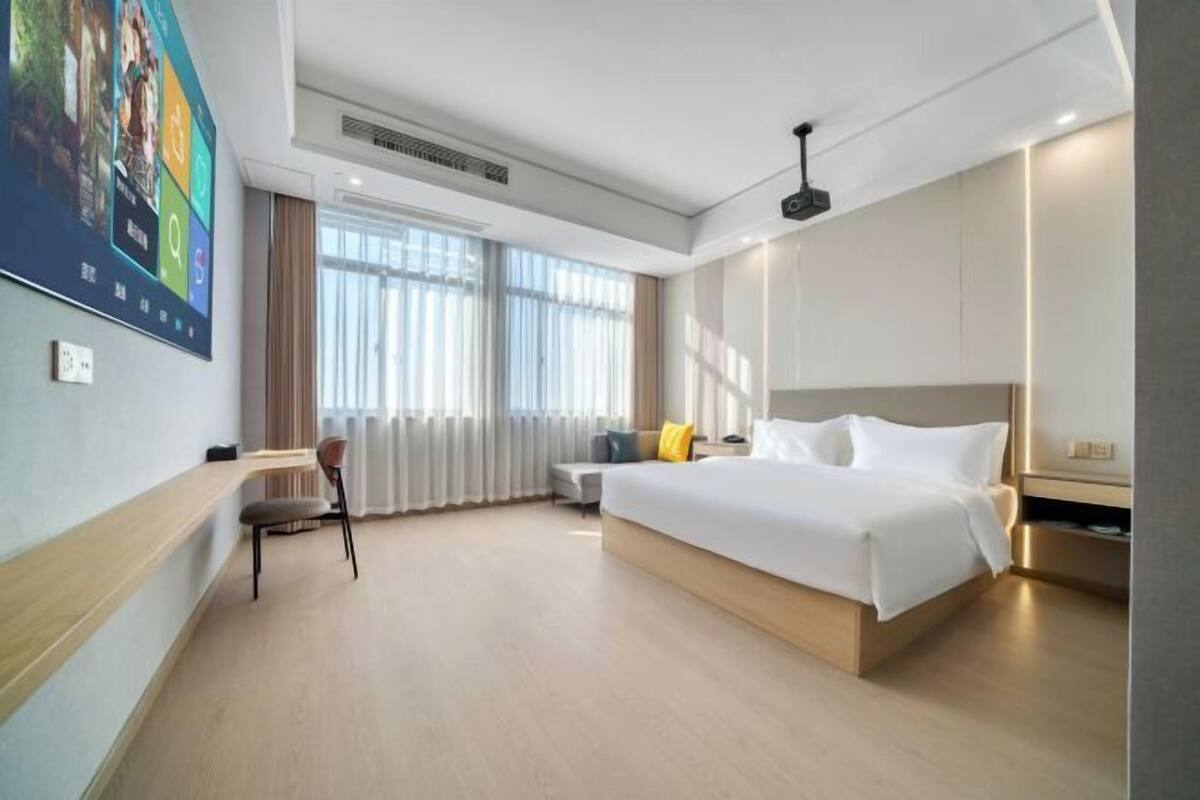 A modern hotel room with a large bed, a desk, and a chair. There is a window with curtains and a ceiling light.