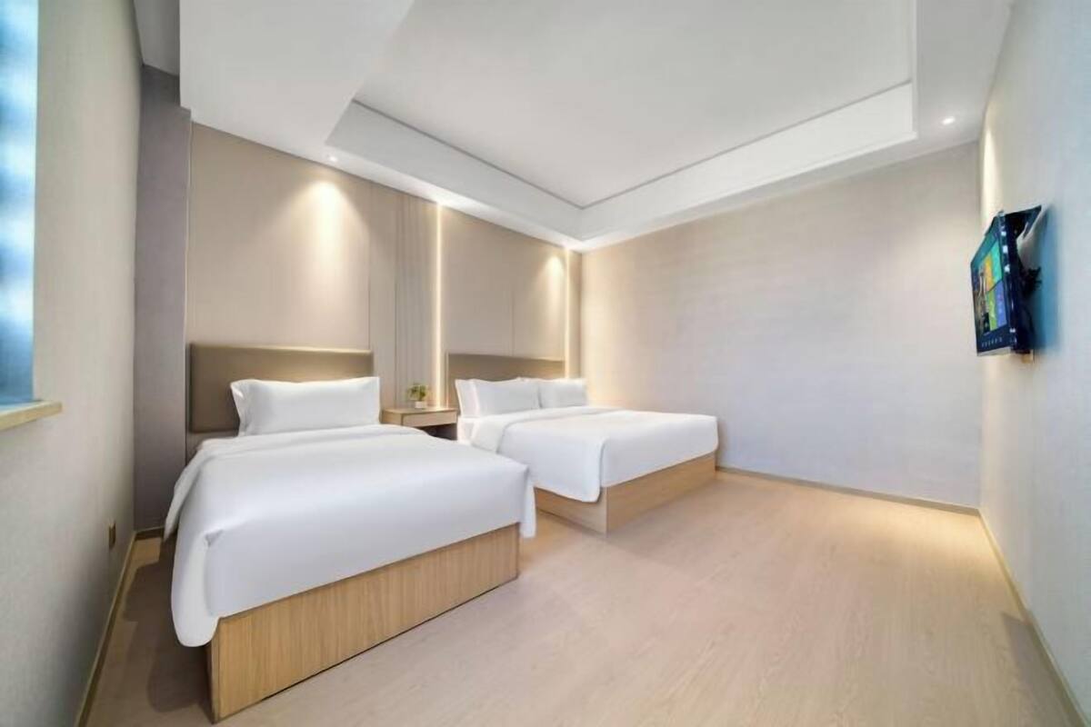 A modern hotel room with two single beds, a small bedside table, a wall-mounted TV, and a large window.