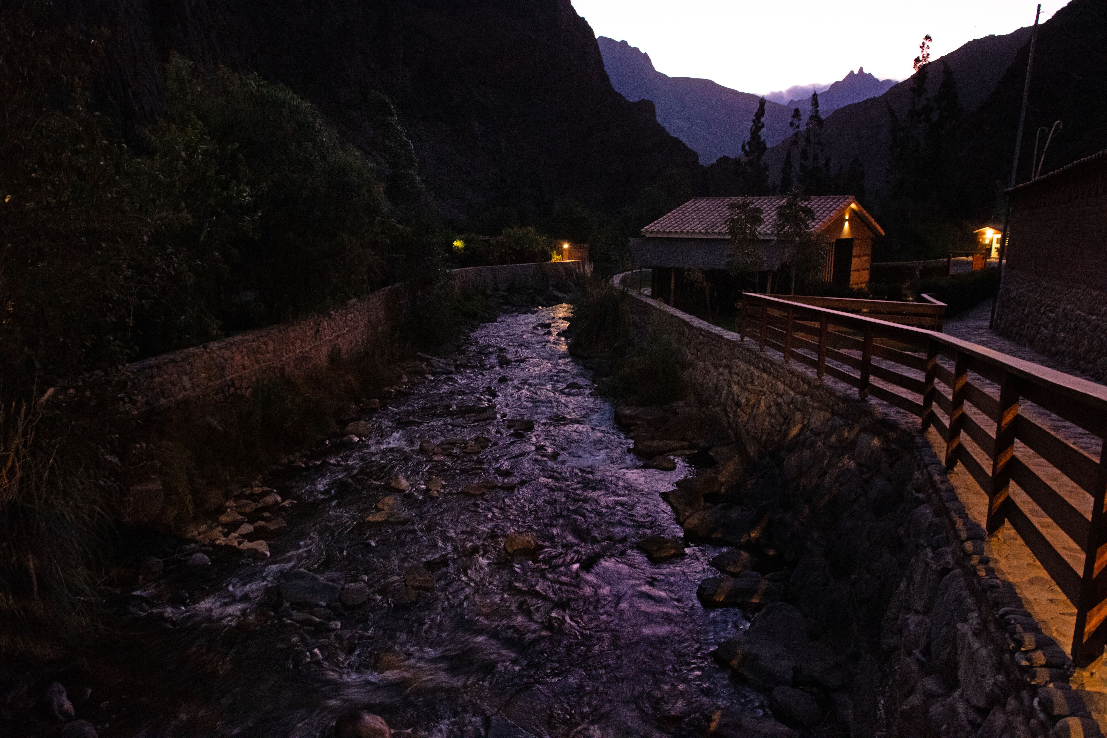 A river flowing through a canyon with a wooden bridge and a small building with lights on.