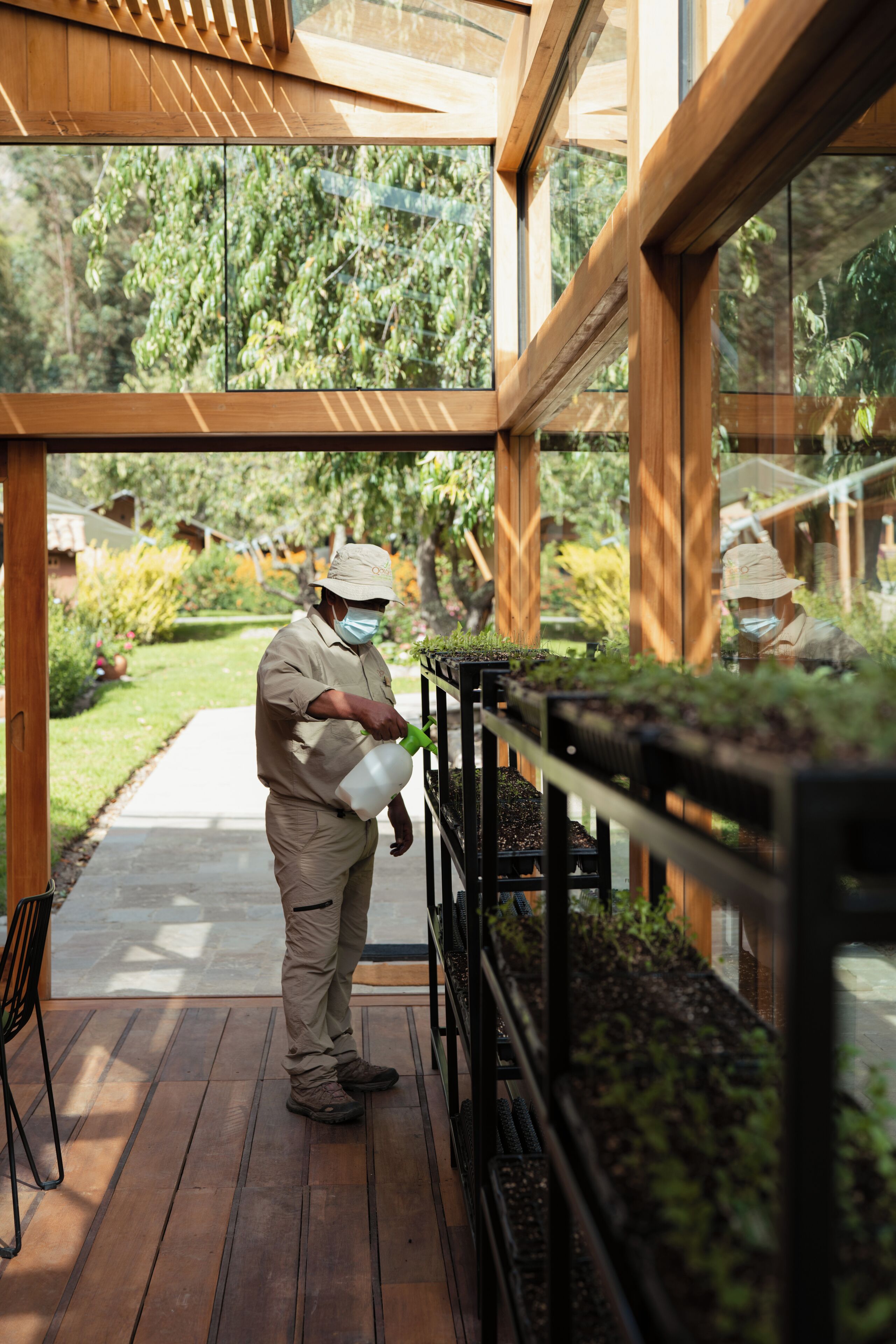 Une personne arrose des plantes à l’intérieur d’une structure en bois aux murs de verre.