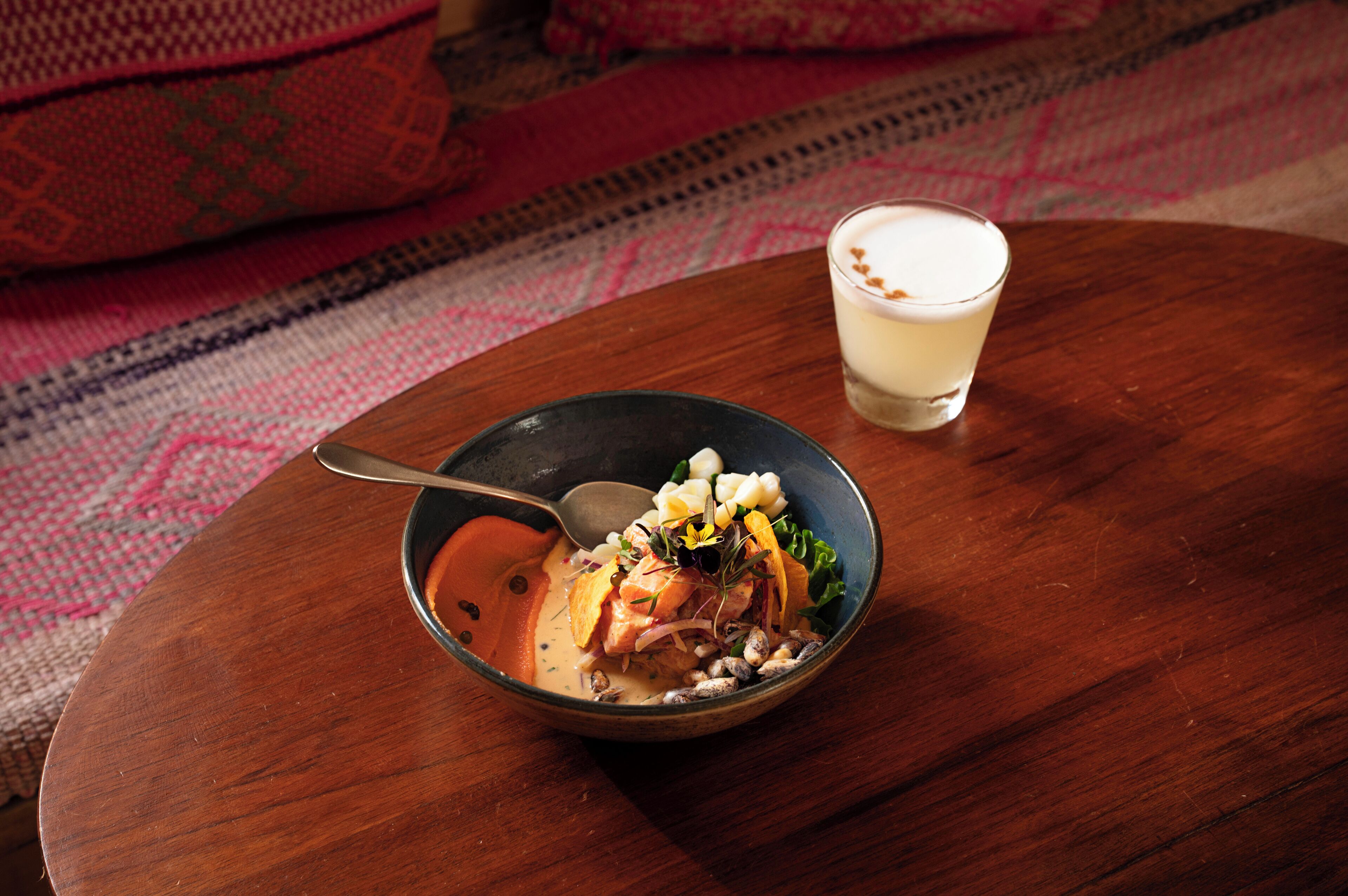 A bowl of food on a wooden table with a spoon, next to a glass of a creamy beverage.