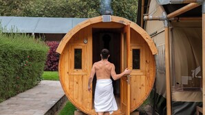 Puinen sauna, josta henkilö on juuri astumassa ulos. Saunan ympärillä on kivetty polku ja vehreää kasvillisuutta.