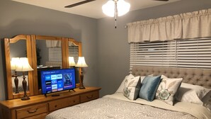 Un dormitorio con cama, televisor sobre un aparador de madera, ventilador de techo y ventana con persianas.