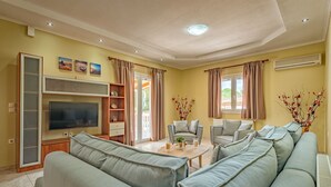 A living room with a large grey sofa, a TV, and a coffee table. There are two armchairs and a bookshelf.
