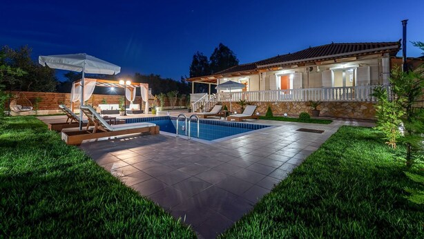 A well-lit outdoor swimming pool area with lounge chairs, a covered seating area, and a stone building with a tiled roof.