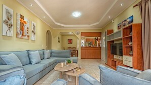 A living room with a sofa, coffee table, and TV stand. There is a kitchen in the background.