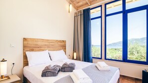 Een slaapkamer met een houten hoofdbord, een groot bed met witte lakens, twee handdoeken en uitzicht op groen door grote ramen.