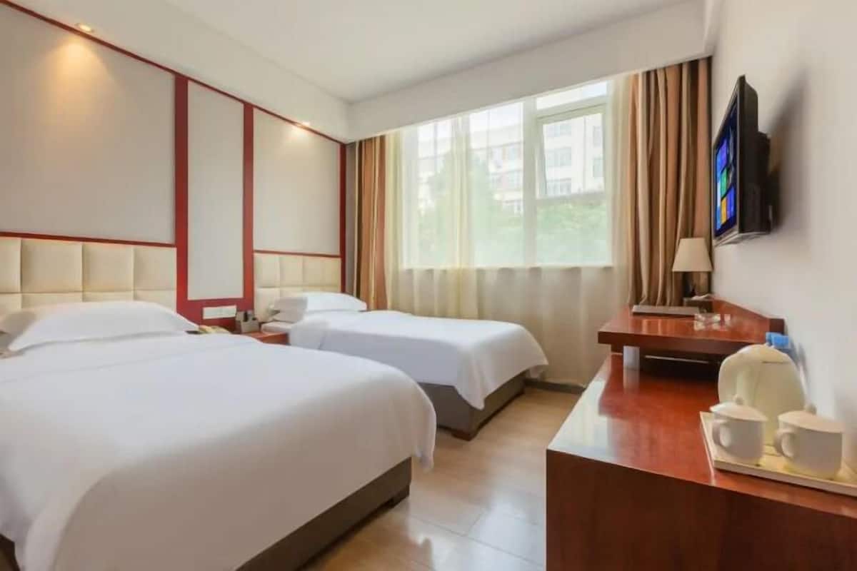 Ein Hotelzimmer mit zwei Betten, einem Schreibtisch mit Lampe, einem Fernseher und einem Fenster mit Vorhängen.