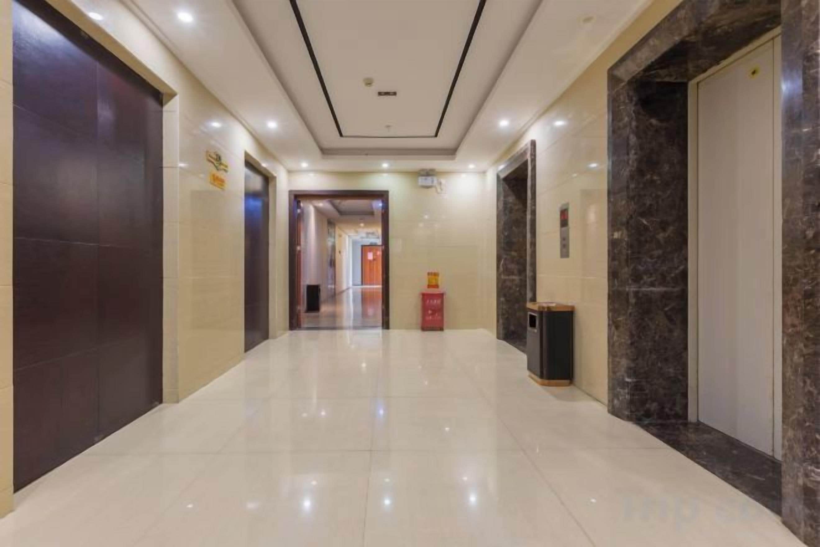 A hotel corridor with a shiny tiled floor, a dark wooden door on the left, a marble wall on the right, and a red trash bin in the center.