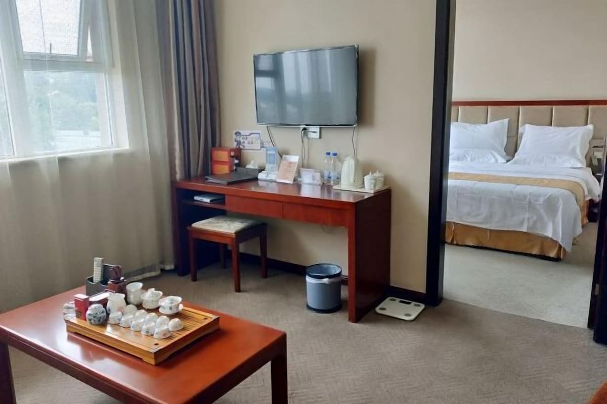 A hotel room with a bed, a desk with a TV, a table with teaware, and a window with curtains.