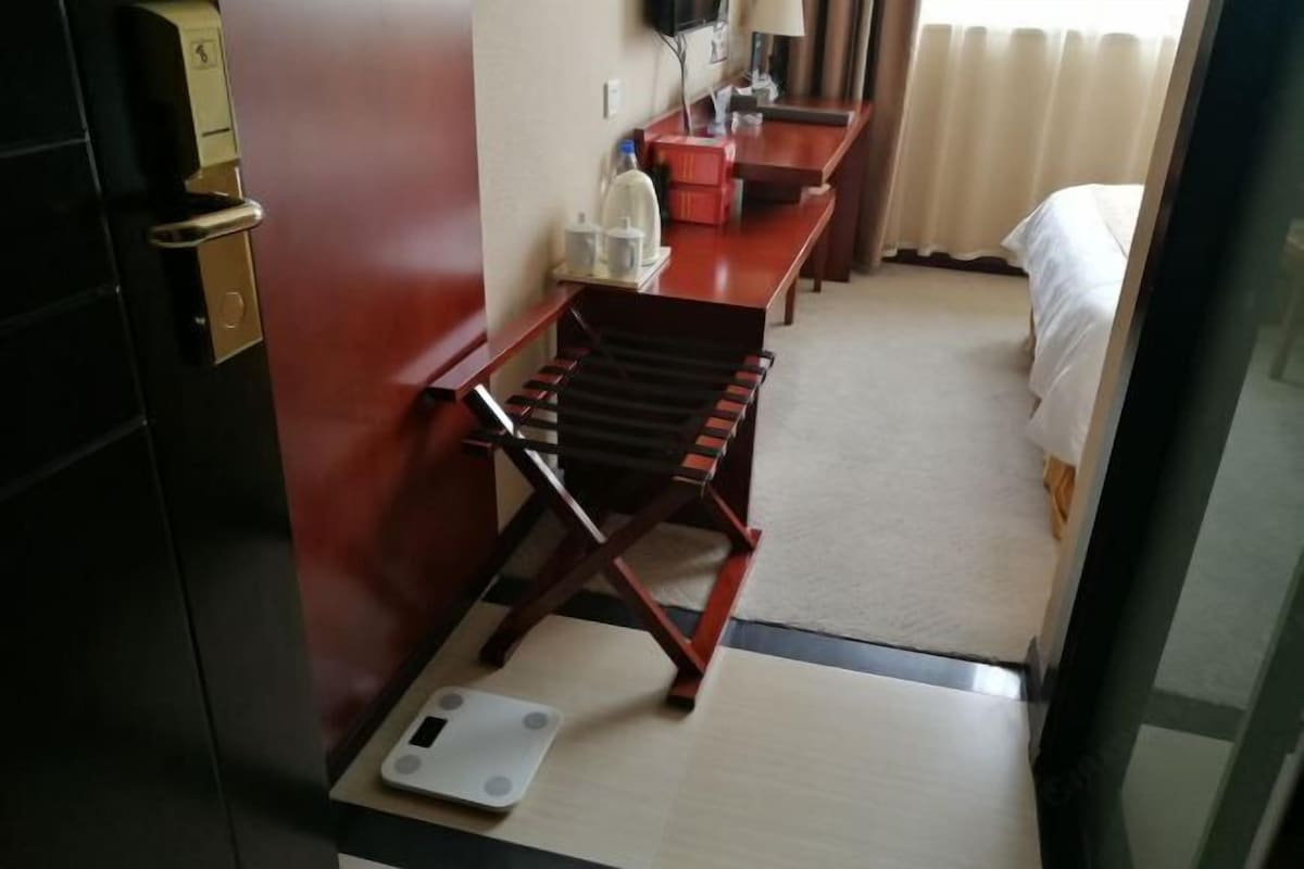 A hotel room with a bed, a desk with a lamp, a chair, a scale, and a red wall.