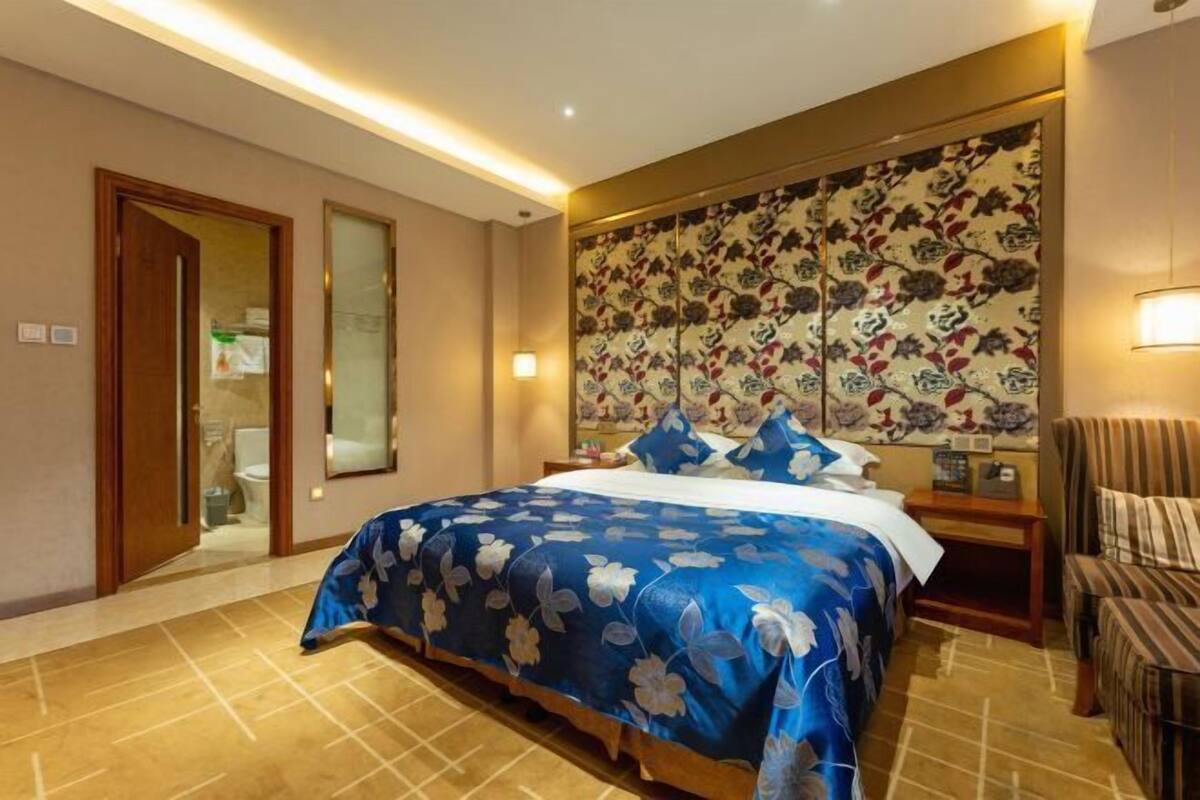 A hotel room with a large bed, a wooden headboard, a nightstand, a chair, and a bathroom visible through an open door.