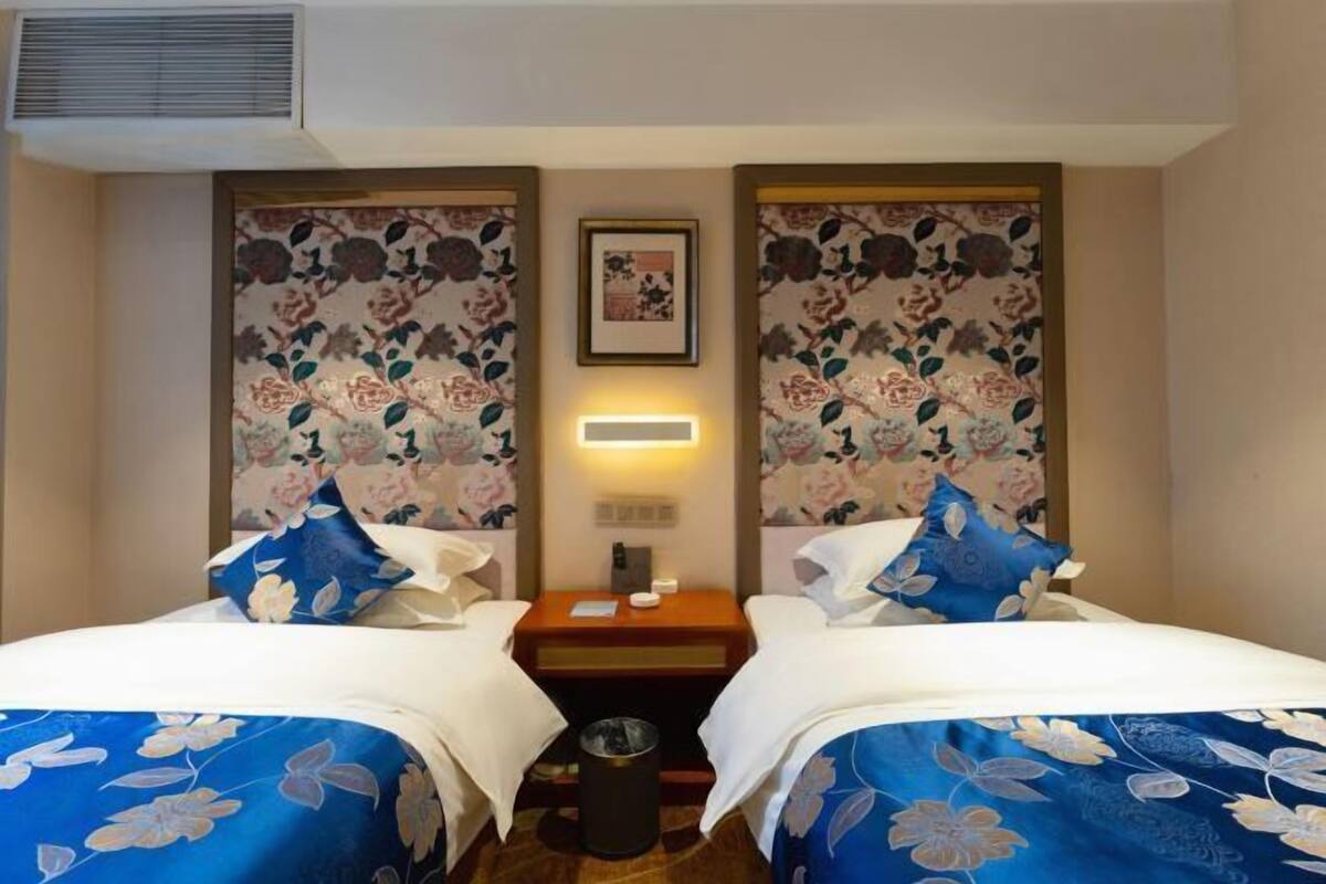 A hotel room with two beds, each with a floral bedspread, a nightstand between them, a wall-mounted light, and a framed picture above the nightstand.