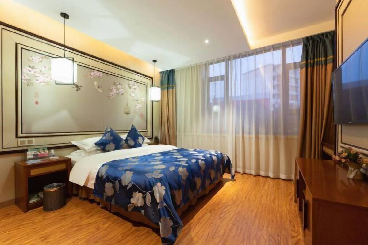 A hotel room with a large bed, wooden flooring, a bedside table with a lamp, a TV, and a large window with curtains.