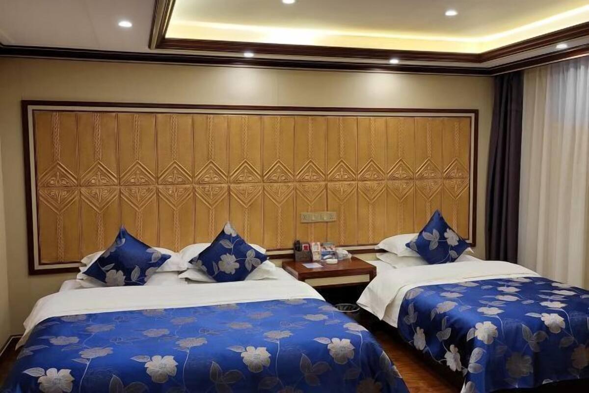 A hotel room with two beds, each with a blue floral bedspread and matching pillows. The room features a large wooden headboard with a geometric pattern and a nightstand with a lamp and a phone.