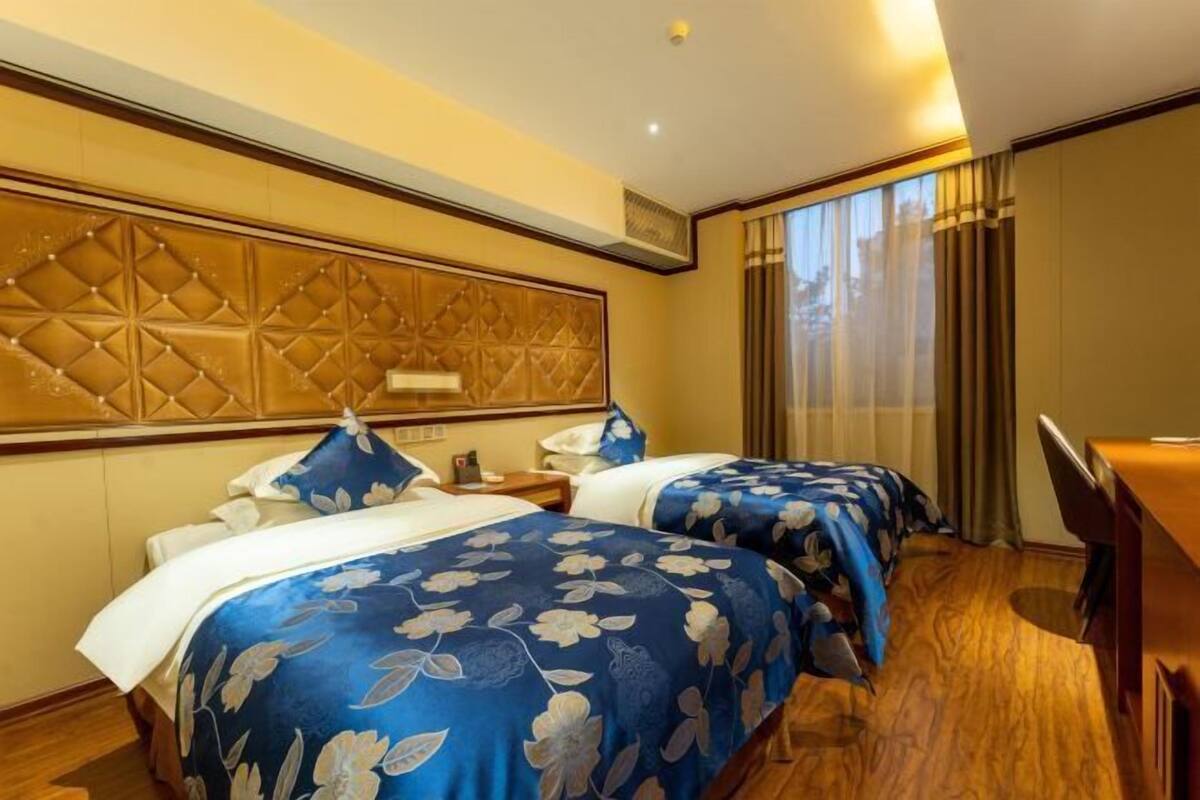 A hotel room with two beds, a wooden headboard, a desk, and a large window with curtains.